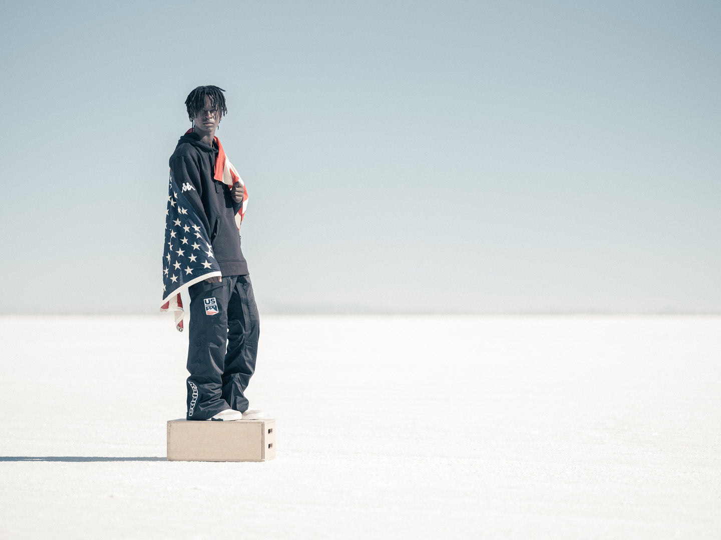 Male US athlete wearing hoodie + ski pants with american flag wrapped around his torso.